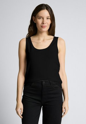 Kobieta z długimi ciemnymi włosami, ubrana w czarny top bez rękawów i czarne spodnie, stojąca na tle jasnoszarym.