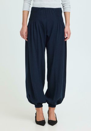 Kvinde iført løstsiddende marineblå bukser med elastisk talje og manchetter, kombineret med sorte spidse hæle og en lysegrå top.