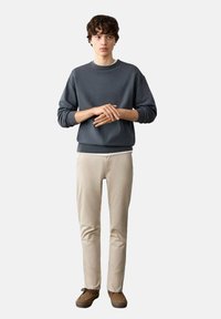 Graues Sweatshirt aus Baumwolle, beige schlanke Hose, braune Schuhe. Das Model steht gerade, die Hände vor dem Körper positioniert, mit lockigem Haar. Minimalistisches Design.
