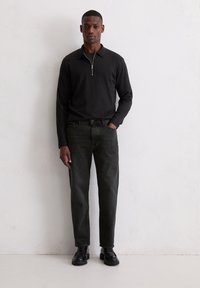 Zwarte poloshirt met lange mouwen en een ritssluiting bij de hals, gecombineerd met donkergroene jeans met rechte pijpen en zwarte veterlaarzen, tegen een lichte achtergrond.