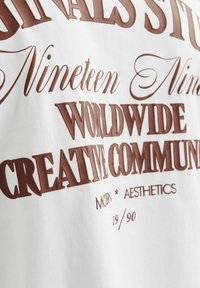 Camiseta blanca con texto impreso en marrón en varios tipos de letra: frases "Diecinueve Noventa," "Comunidad Creativa Global," y "Estética Moderna."