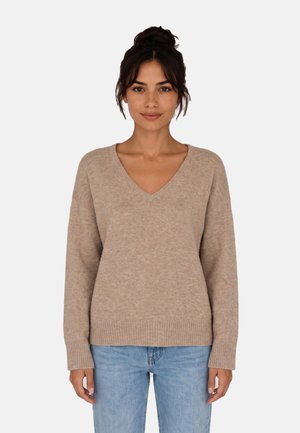 Donna con capelli scuri raccolti in uno chignon, che indossa un maglione beige con scollo a V e jeans azzurri chiari, in piedi davanti a uno sfondo bianco semplice.