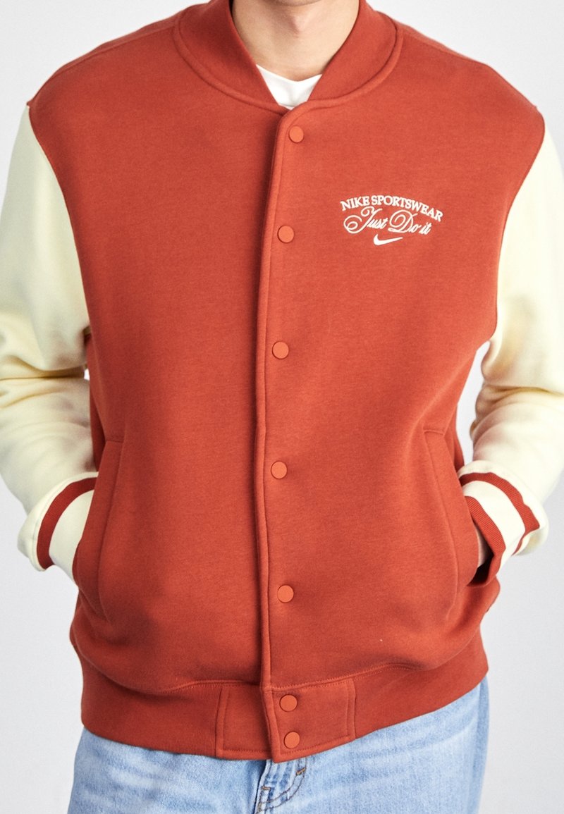 Nike sportjacka i rostorange med cremefärgade ärmar. Har tryckknappar, logobrodyr och ribbade detaljer vid manschetter och krage.