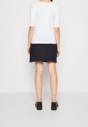 Femme debout, dos tourné, portant un haut blanc à manches courtes, une jupe noire avec des poches à l’arrière et des chaussures noires sur un fond uni.