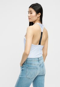 Γυναίκα με μακριά σκουρόχρωμα μαλλιά, που φορά λευκό αμάνικο τοπ με μαύρο λουρί στο πίσω μέρος και ανοιχτό μπλε τζιν, στέκεται με τα χέρια στις τσέπες.