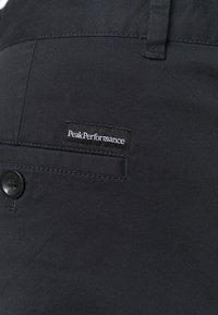 Detailní záběr na kapsu černých látkových kalhot s knoflíkem a označením "Peak Performance" našitým nad kapsou.