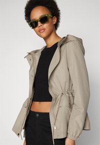 Lys beige, glidelåsjakke med hette, innsnørt liv og elastiske mansjetter. Bært over en svart strikket topp, parret med transparente grønne solbriller.