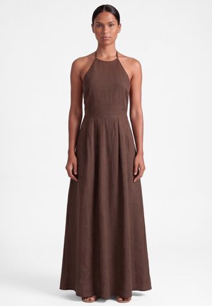 Kobieta w brązowej, bezrękawnej sukni na halter, sięgającej ziemi, stojąca na jednolitym białym tle.