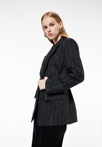 Raitis mustapuku, jossa on pystysuorat raidat, varustettu läpällä, kahdella etutaskulla ja yhdellä napin kiinnityksellä. Hienovarainen kankaan vaihtelu.