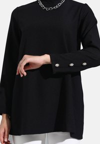 Maglietta nera a maniche lunghe in tessuto liscio, con colletto a lupetto, collana di catena metallica e bottoni decorati sulle maniche.