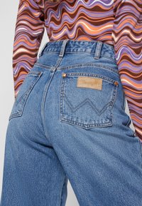 Korkeavyötäröiset siniset farkut, joissa on tekstuuroitu pinta, kaksi takataskua, nahkainen logopatch ja kontrastivärisiä ompeleita.