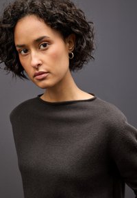 Dark sweater with a high, wide neckline, featuring a soft, textured knit. Model has curly hair and gold hoop earrings, against a neutral background.