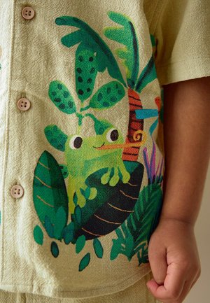 Béžová košile s ilustrací zelené žáby a tropické rostliny, dřevěné knoflíky a pravá ruka osoby opřená vedle košile.