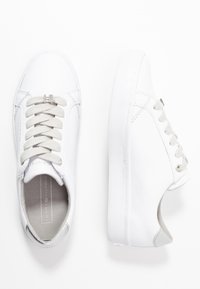 White low-top sneakers with beige laces, silver lace charm, and Tommy Hilfiger branding on the insole and back heel.