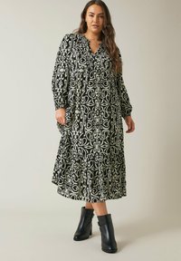 Schwarz-weiß gemustertes Kleid mit einem gestuften Rock, V-Ausschnitt und langen Ärmeln. Kombiniert mit schwarzen Stiefeletten.