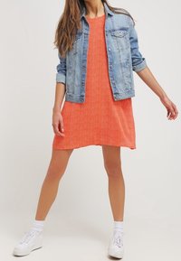 Veste en jean portée sur une robe orange à motif zigzag ; le mannequin porte des baskets blanches et des chaussettes courtes, posant debout.