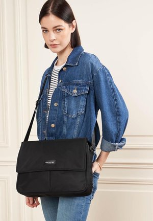 Zwarte nylon schoudertas met een klep sluiting, voorzien van een merketiket, gedragen als crossbody. Gecombineerd met een blauwe denimjack en een gestreept shirt.