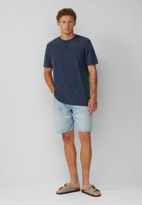 Navyblaue Baumwoll-T-Shirt, kurze Ärmel, runder Ausschnitt. Hellblaue Jeansshorts mit ausgefranstem Saum. Braune Sandalen mit zwei Riemen und einer Schnalle.