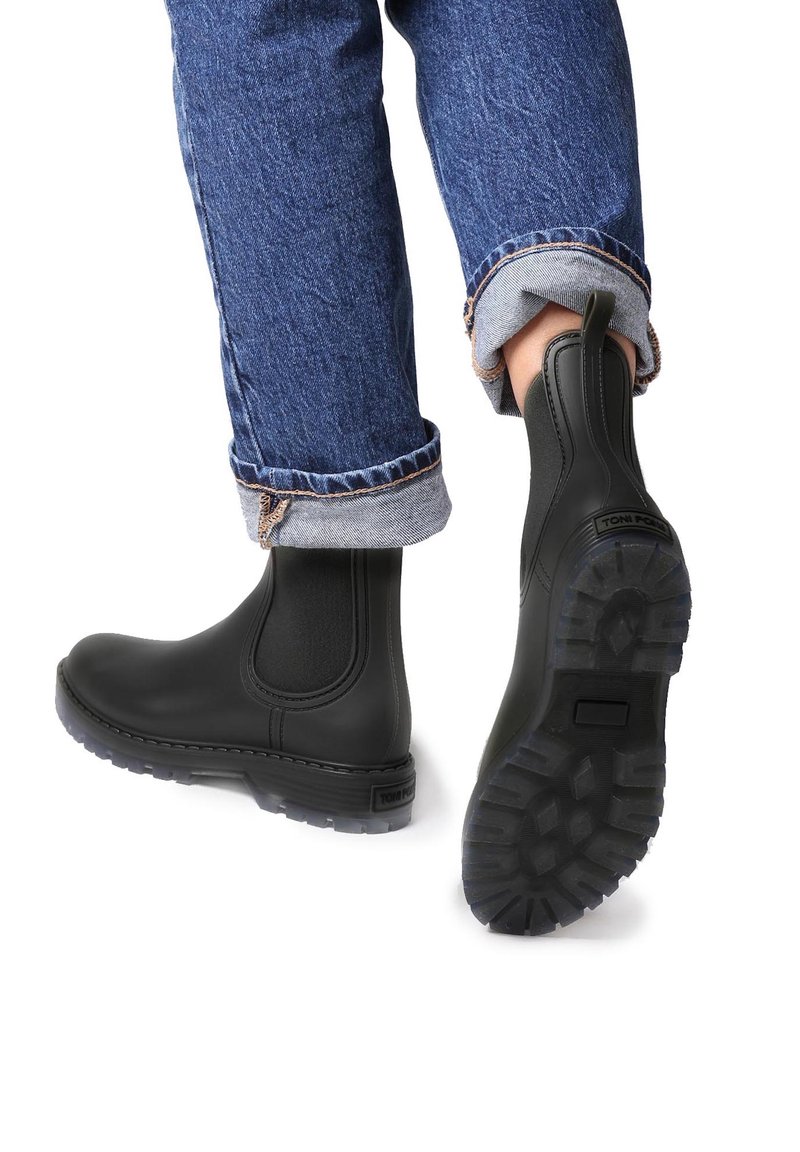 Personne portant des bottines Chelsea noires à semelles crantées et un jean bleu retroussé, un pied levé montrant le motif de la semelle de la bottine.