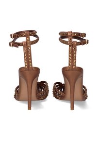 Sandalias de tacón alto marrones con puntera abierta, que presentan remaches decorativos dorados en las correas del tobillo y un diseño entrelazado. Textura de cuero suave.