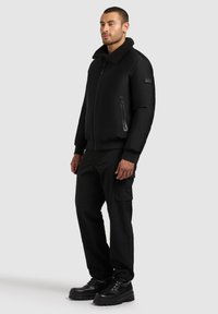 Jachetă neagră din bomber cu guler din blană de oaie, buzunare cu fermoar și manșete ribbate, asortată cu pantaloni cargo negri și cizme negre rezistente.