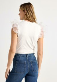 Hvid top med gennemsigtige flæseærmer, kombineret med mørkeblå, højtaljede jeans med baglommer og klassisk denimstruktur.