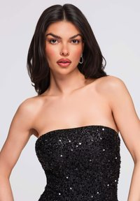Robe noire sans bretelles recouverte de sequins, avec un corsage ajusté et une texture lisse. Accessoirisée de boucles d'oreilles étincelantes.