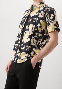 Camisa de manga corta con un patrón floral en amarillo y negro, confeccionada en tela suave, con cuello puntiagudo y cierre de botones.