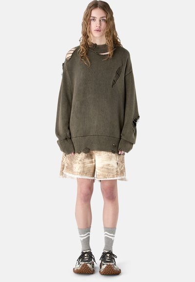 Młoda osoba z długimi włosami, ubrana w za dużą, podartą ciemnooliwkową sweter, beżowe szorty, szare skarpetki i czarno-białe trampki, stoi przodem.