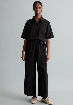 Jeune femme debout de face, portant une chemise noire ample à manches courtes boutonnée, un pantalon large noir et des chaussures beige avec des lacets roses.
