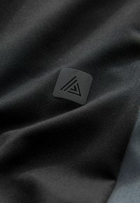 Una tela negra con una textura suave presenta un pequeño logotipo cuadrado con un diseño geométrico, acentuado por una superficie negra elevada.
