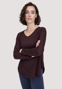 Frau mit kurzem, lockigem, dunklem Haar, die ein langärmliges, dunkelpurpurnes Oberteil und eine marineblaue Hose trägt und mit verschränkten Armen vor einem einfarbigen Hintergrund steht.