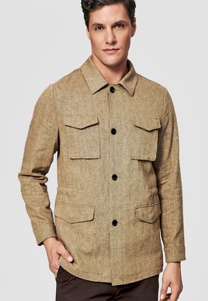 Mężczyzna ubrany w beżową, zapinaną na guziki kurtkę z czterema przednimi kieszeniami z klapami oraz ciemne spodnie, stojący na jednolitym jasnym tle.