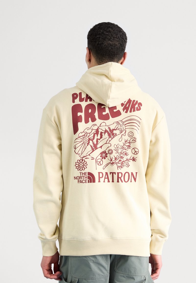 Homme portant un sweat à capuche crème avec un texte graphique rouge "Plant Freaks" et des illustrations de main, de fleurs, de signes de paix et de montagnes au dos.