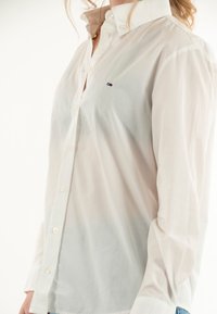 Camicia bianca con bottoni, realizzata in tessuto leggero, caratterizzata da un colletto a punta, un logo sul petto e bottoni in plastica lungo la chiusura.