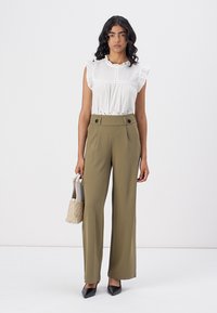 Blusa bianca a ruffles con dettagli in pizzo, pantaloni a palazzo verdi oliva, décolleté nere a punta e una piccola borsa beige testurizzata.