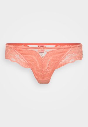 String en dentelle corail avec des bords festonnés, un nœud central et une taille en satin lisse. Présente des motifs floraux complexes et un dos minimaliste.