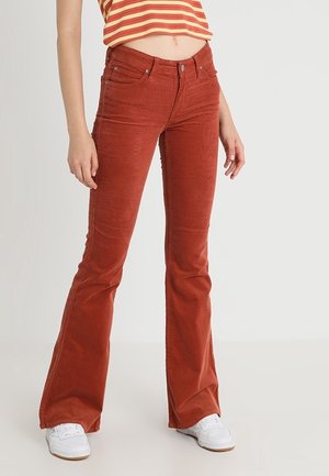 Kvinde iført rustrøde flarebukser, stribet cropped top og hvide sneakers, stående mod en ensfarvet lys baggrund.