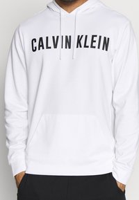 Sweat à capuche blanc en mélange de coton, avec une poche avant et un texte noir audacieux "CALVIN KLEIN" sur la poitrine.