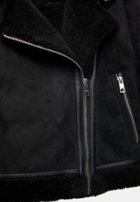 Veste en shearling noire à finition texturée, avec un grand col, une fermeture éclair argentée et des poches latérales ornées de détails métalliques élégants.