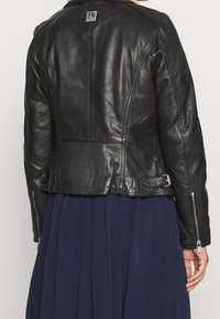 Giacca in pelle nera con un design aderente, caratterizzata da un colletto alto, accenti metallici e polsini con zip. Texture liscia con cuciture strutturate.