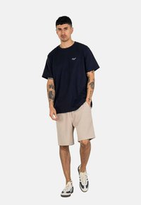 Camiseta de algodón azul marino de manga corta con un pequeño logo, combinada con pantalones cortos beige y zapatillas blancas con detalles azules.