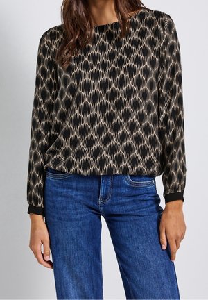 Kvinde iført en langærmet bluse med sort og beige abstrakt mønster og blå jeans, står mod en ensfarvet lys baggrund.