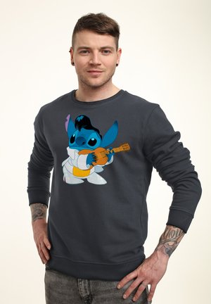 Man met tatoeages draagt een donker sweatshirt met daarop een cartoonfiguur die gitaar speelt, staand met de handen in de zij tegen een effen achtergrond.