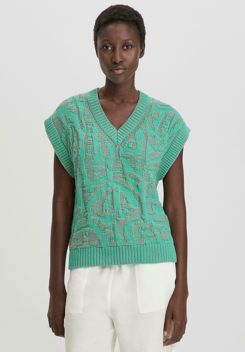 Grüner, gestrickter ärmelloser Pullover mit V-Ausschnitt, der ein strukturiertes Muster und einen gerippten Saum aufweist. Kombiniert mit hellen, locker sitzenden Hosen.