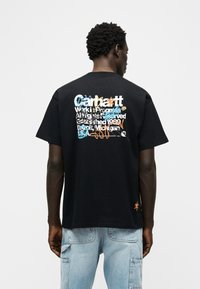 Άνδρας που φοράει μαύρο μπλουζάκι με πολύχρωμο γραφίτι στυλ κείμενο στην πλάτη και ανοιχτό μπλε τζιν με τσέπες cargo.