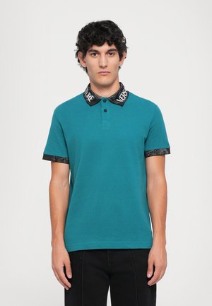 Bărbat cu păr curly, îmbrăcat cu un tricou polo teal, cu guler și manșete negre cu model, stând în fața unui fundal alb simplu.