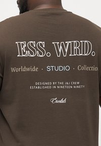 Brun bomulds t-shirt med hvid og lyseblå teksttryk på ryggen, med designdetaljer der fremhæver mærket og kollektionen.