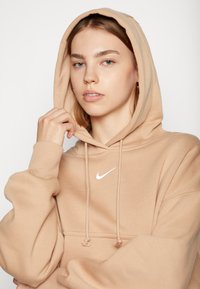 Sweatshirt bege com cordão e bolso canguru, apresentando um logótipo branco da Nike no peito e punhos passantes. Textura de tecido suave.