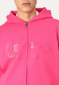 Ružová mikina na zips s embosovaným logom „Colucci la Famiglia“, vyrobená z mäkkej látky s kapucňou na šnurovanie a kovovým zipsom.